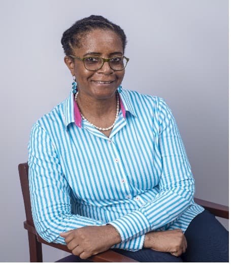 Dr Ayo harriman dressed in a blue and white pin striped collared shirt, while seated in an armchair.