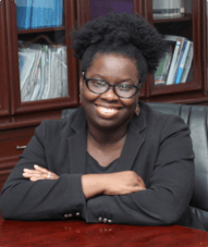 Professional portrait of Dr Feyi Hassan-Oyewumi wearing a black blazer and matching blouse while seated at mahogany table.